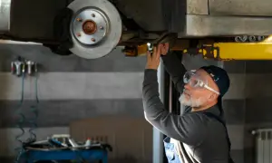 Mechanik w okularach ochronnych pracuje pod samochodem, być może szukając sposobu, jak odblokować hamulec ręczny elektryczny.
