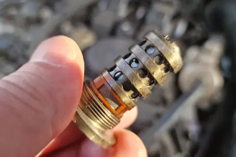 Uszkodzony zawór zmiennych faz rozrządu objawy: brudny, metalowy element z otworami, trzymany w palcach.