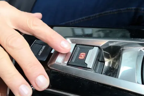 Dłoń naciska przycisk hamulca ręcznego. Tak zaciągnąć hamulec ręczny w nowoczesnym samochodzie.