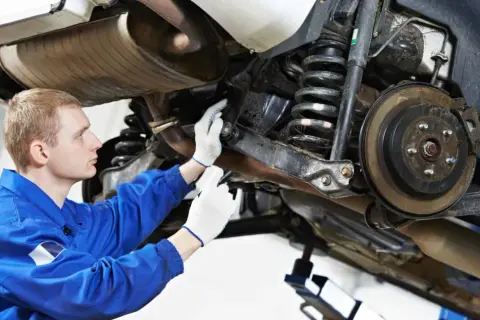 Mechanik w niebieskim kombinezonie sprawdza elementy zawieszenia samochodu, w tym sprężyny i hamulce.