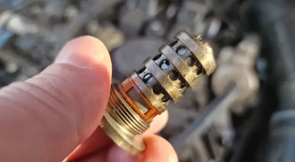 Uszkodzony zawór zmiennych faz rozrządu objawy: brudny, metalowy element z otworami, trzymany w palcach.