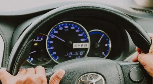 Dłonie na kierownicy Toyoty, widoczne zegary. Trudności ze skręcaniem mogą oznaczać zapowietrzony układ wspomagania objawy.