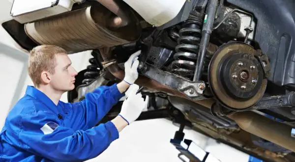 Mechanik w niebieskim kombinezonie sprawdza elementy zawieszenia samochodu, w tym sprężyny i hamulce.