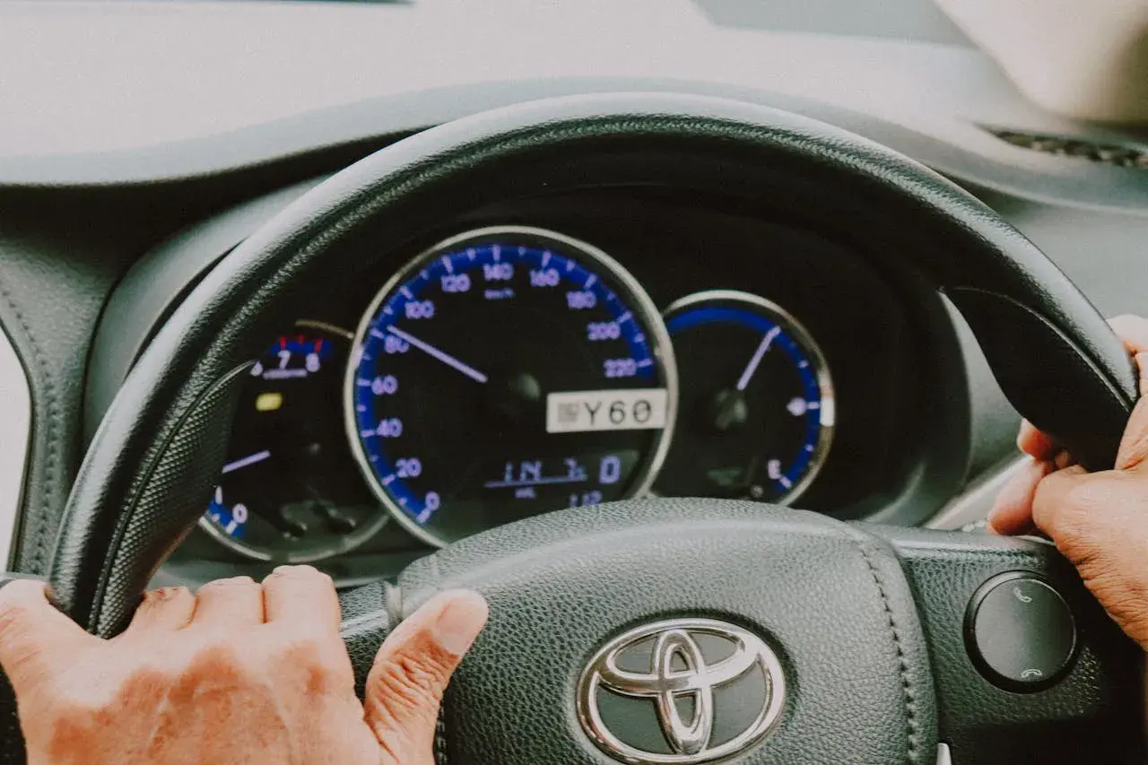 Dłonie na kierownicy Toyoty, widoczne zegary. Trudności ze skręcaniem mogą oznaczać zapowietrzony układ wspomagania objawy.