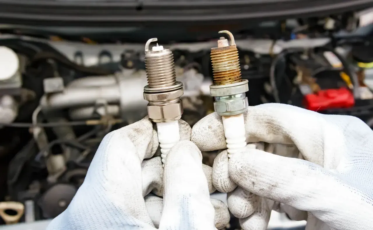 Ręce w białych rękawiczkach trzymają dwie świece zapłonowe, jedną nową, drugą zużytą. Czy diesel ma świece zapłonowe? Nie, diesel nie ma świec zapłonowych.