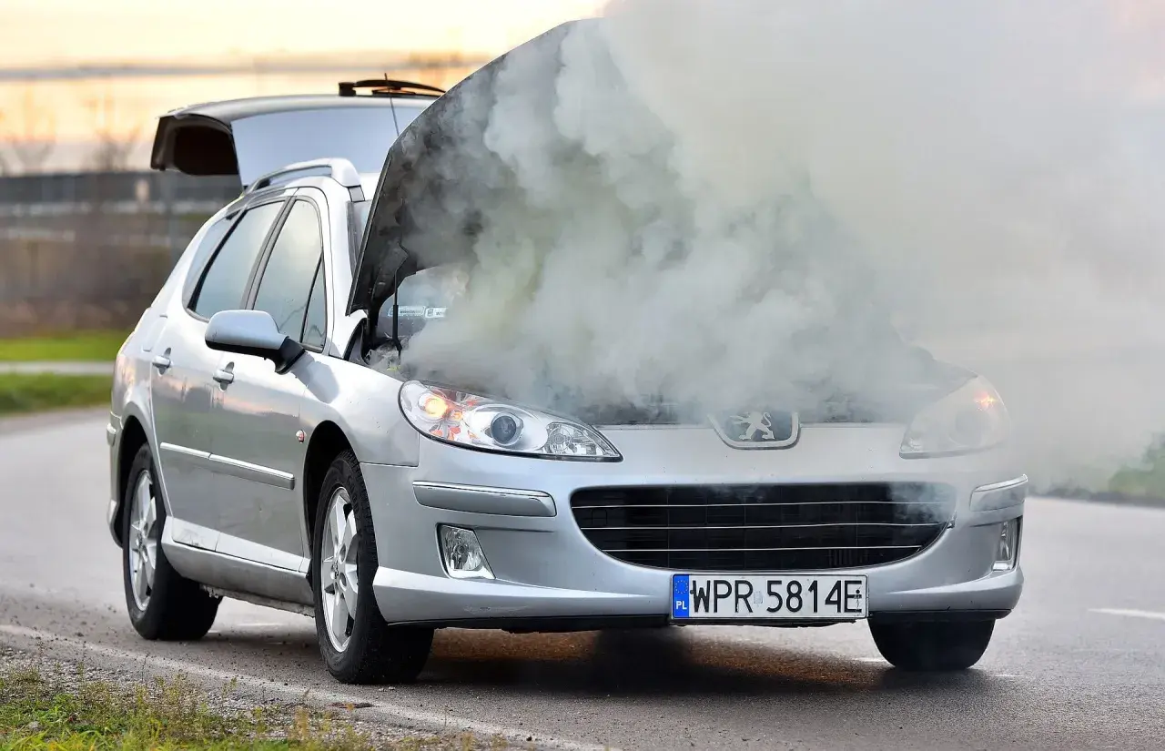 Srebrny Peugeot z otwartą maską i dymem wydobywającym się z silnika. Rozbiegany silnik diesla na poboczu drogi.