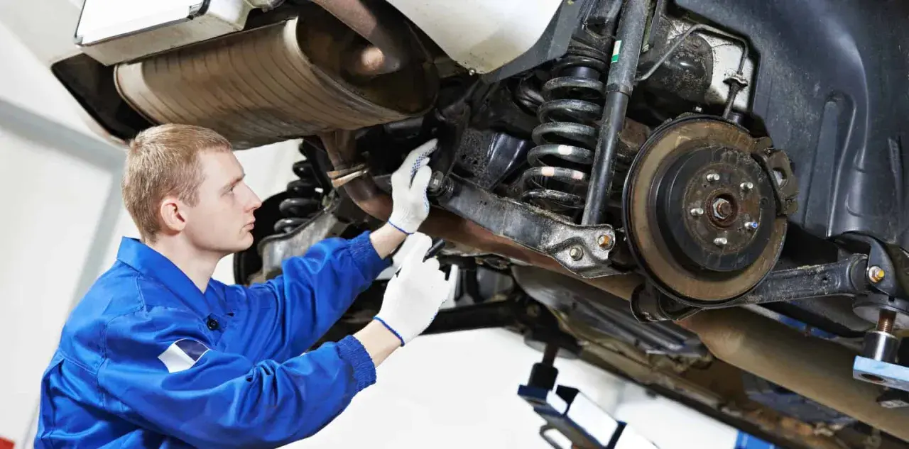 Mechanik w niebieskim kombinezonie sprawdza elementy zawieszenia samochodu, w tym sprężyny i hamulce.