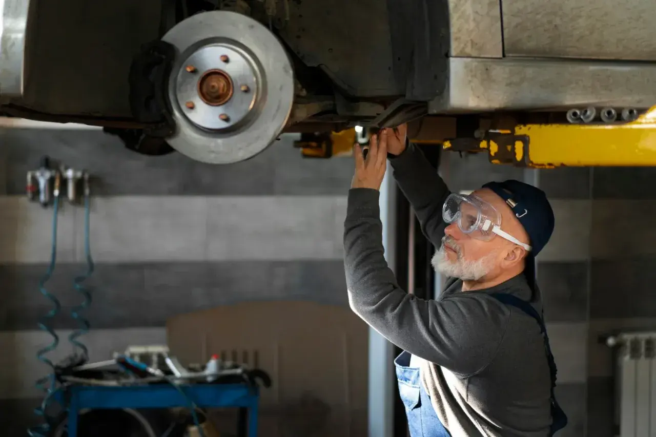 Mechanik w okularach ochronnych pracuje pod samochodem, być może szukając sposobu, jak odblokować hamulec ręczny elektryczny.