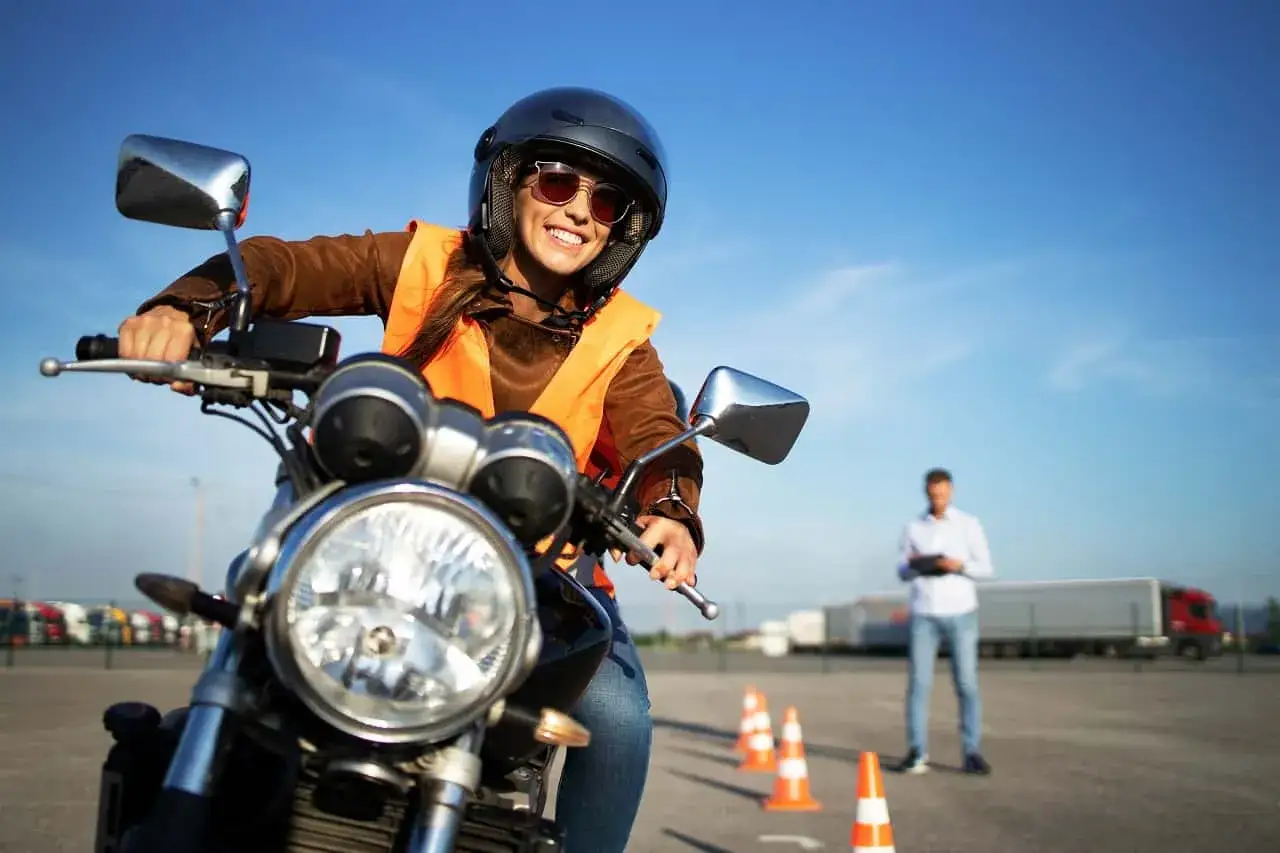 Uśmiechnięta kobieta w kasku i kamizelce na motorze, gotowa do zdobycia prawa jazdy na motor. Instruktor obserwuje z tyłu.