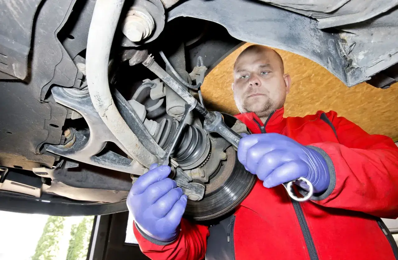 Mechanik w niebieskich rękawiczkach dokonuje wymiany końcówki drążka kierowniczego, używając kluczy do precyzyjnego dokręcenia.